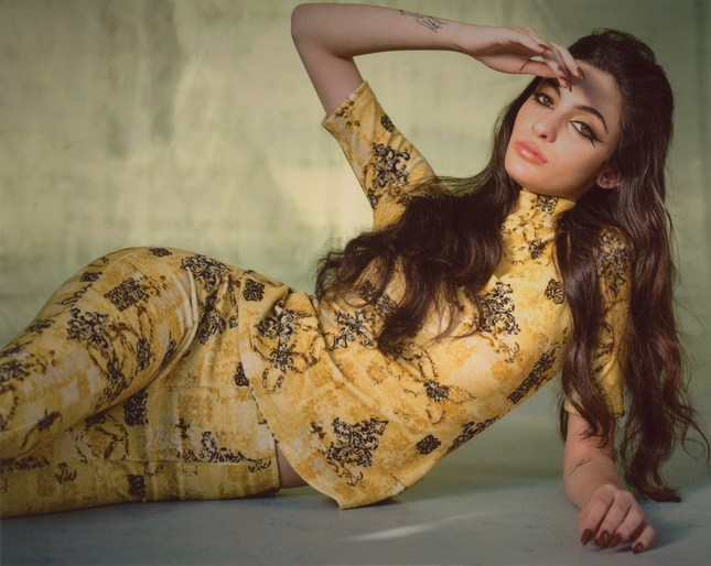 Woman in a patterned top and pants poses reclining in a studio with her hand shielding her eyes, displaying a fashion-forward look.