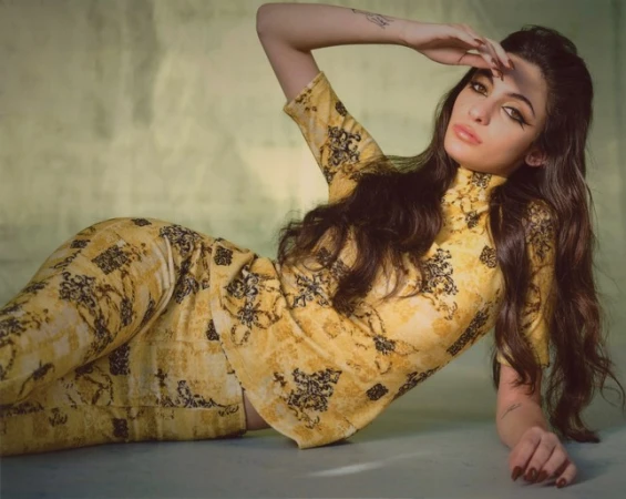 Woman in a patterned top and pants poses reclining in a studio with her hand shielding her eyes, displaying a fashion-forward look.