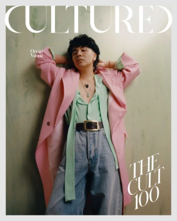 Magazine cover featuring a person with dark hair wearing a pink coat, light-green shirt, blue jeans and a belt, posing in front of a wall.