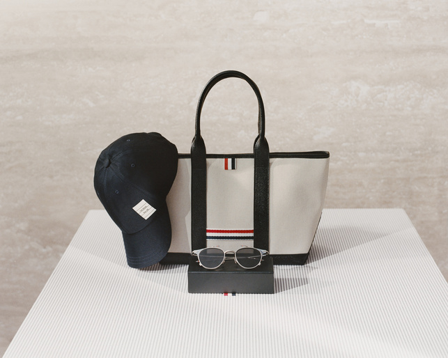 Still life of a tote bag, sunglasses, and a baseball cap, all accented with red, white, and blue stripes, on a striped white surface.