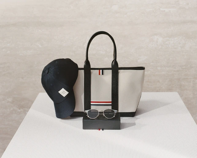 Still life of a tote bag, sunglasses, and a baseball cap, all accented with red, white, and blue stripes, on a striped white surface.
