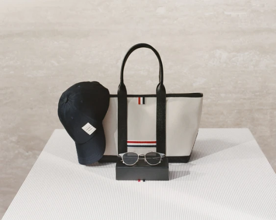 Still life of a tote bag, sunglasses, and a baseball cap, all accented with red, white, and blue stripes, on a striped white surface.