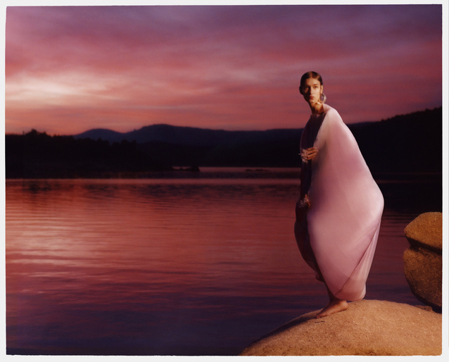 Model standing on a rock by a tranquil lake at dusk wearing a flowing pink dress against a dramatic sunset sky