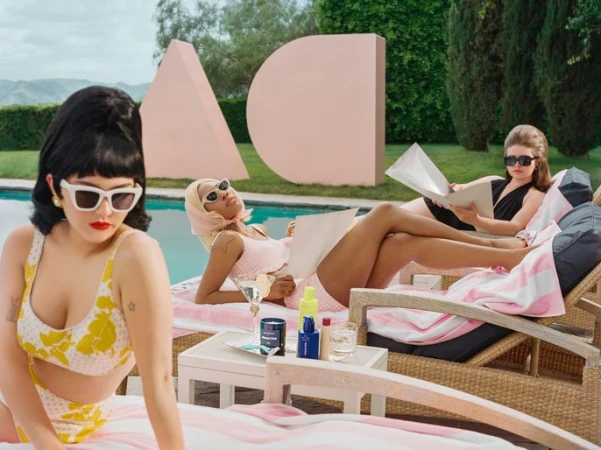 Three women lounge poolside in retro swimwear and sunglasses, reading and enjoying drinks, with geometric art in the background.