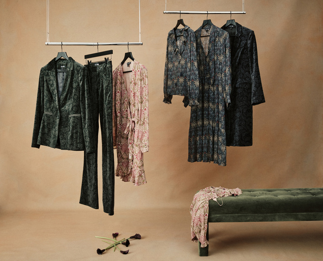 Several pieces of patterned women's clothing hang from racks next to a velvet bench with calla lilies in a warm-toned indoor setting.