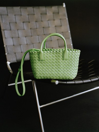 A light green woven Bottega Veneta handbag with handles and a strap sits on a black chair with a woven grey seat against a dark background.