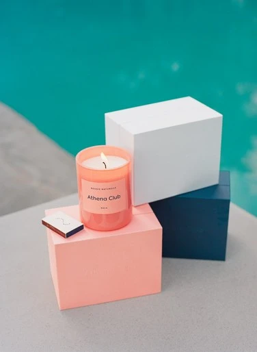 A lit candle sits on a pink box, next to matches, with a blue box and white box in the background against a blue pool.