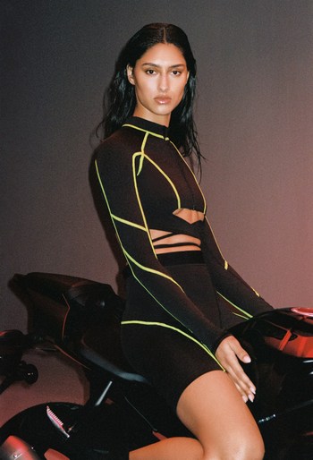 A woman in a black and neon green outfit poses on a motorcycle in an indoor studio with a dark background, in a fashion forward image.