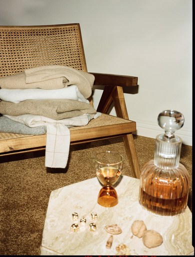 Still life shot of a chair with folded sweaters, glasses, and a decanter on a small marble table against a plain neutral wall.