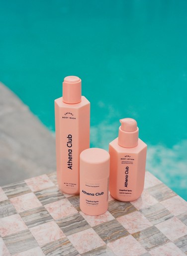 Three pink Athena Club beauty products on a checkered surface with a turquoise pool in the background, showcasing product photography.