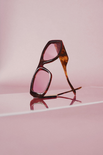 Tortoiseshell sunglasses with pink lenses are angled and reflected against a glossy pink background. The glasses appear to be standing on their arms.