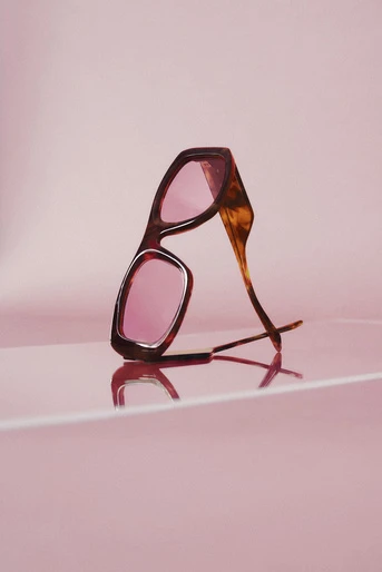 Tortoiseshell sunglasses with pink lenses are angled and reflected against a glossy pink background. The glasses appear to be standing on their arms.