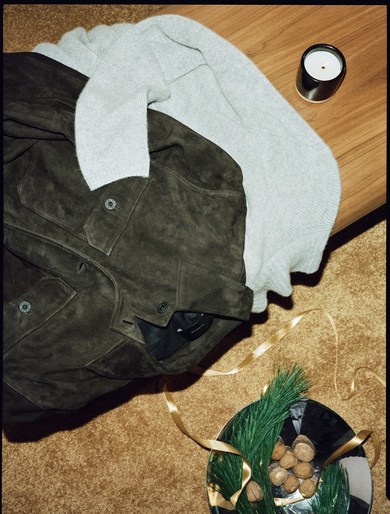 Overhead shot of brown suede jacket, gray sweater, candle, nuts, and pine needles on a brown carpet create a cozy, inviting scene.