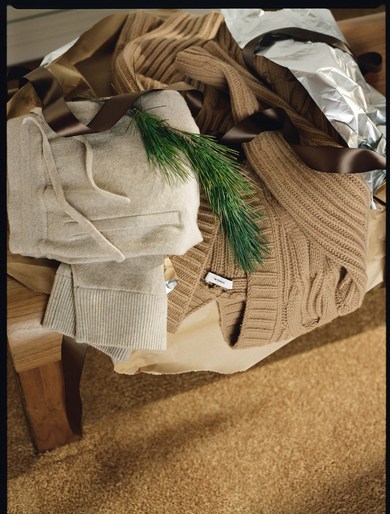 Still life of neatly folded beige knitwear displayed on a wooden surface with a sprig of greenery, brown ribbon, and textured background.