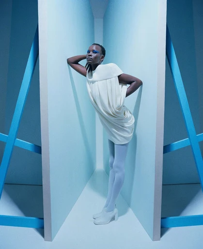 Fashion shot of a slender Black female model in a white dress and white tights leaning against a blue and white structure in a blue room.