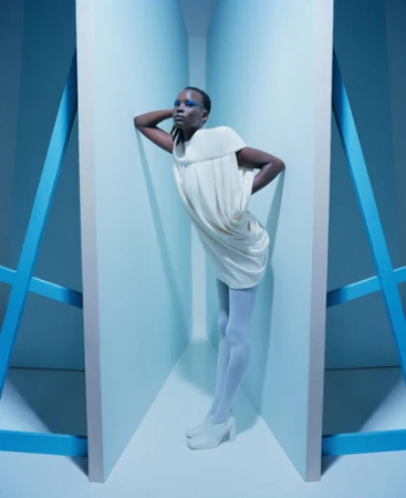 Fashion shot of a slender Black female model in a white dress and white tights leaning against a blue and white structure in a blue room.
