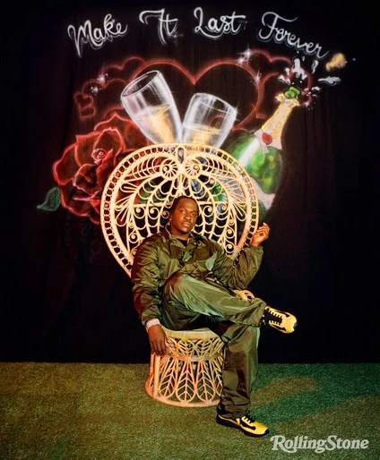 Man in olive-green jumpsuit sits in peacock chair against a backdrop of painted champagne, hearts, and roses, with 'Rolling Stone' logo visible.