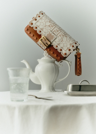 Brown leather Fendi handbag with white lace detail on a white teapot sitting on a table with a glass, fork, and silver butter dish.