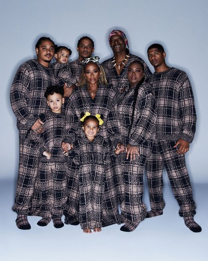 A large group of ten people, including adults and children, pose in matching brown-and-black plaid pajamas against a light blue background.