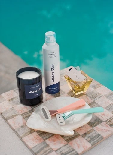 Still life of Athena Club shaving products including razors, shaving cream and body wash on a tile surface next to a pool.