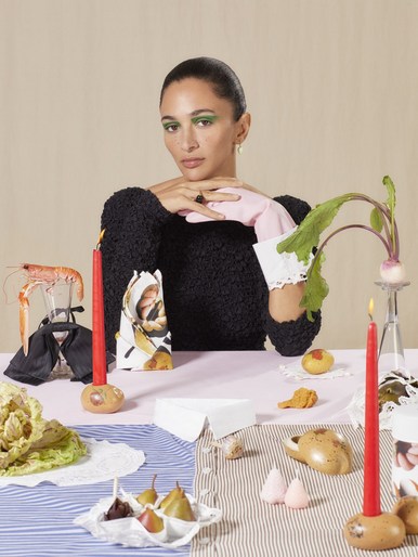 A woman with green eye makeup wearing a black textured sweater and pink gloves sits at a styled table with candles and food.