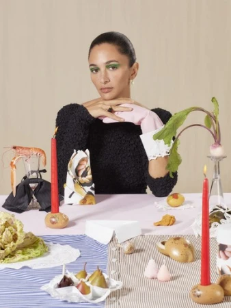A woman with green eye makeup wearing a black textured sweater and pink gloves sits at a styled table with candles and food.