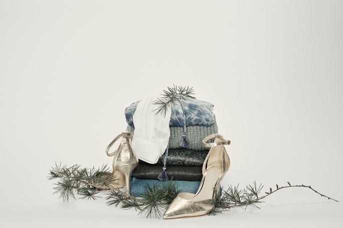 Stack of folded blue, white, and dark-colored fabrics, accessorized with gold high heels and evergreen branches on a white backdrop.
