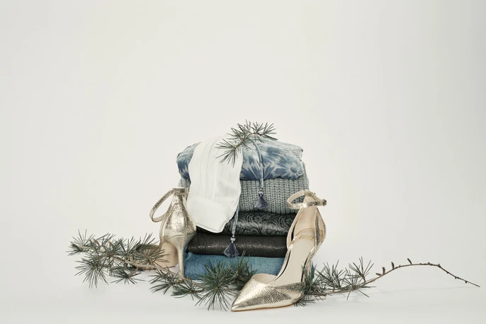 Stack of folded blue, white, and dark-colored fabrics, accessorized with gold high heels and evergreen branches on a white backdrop.