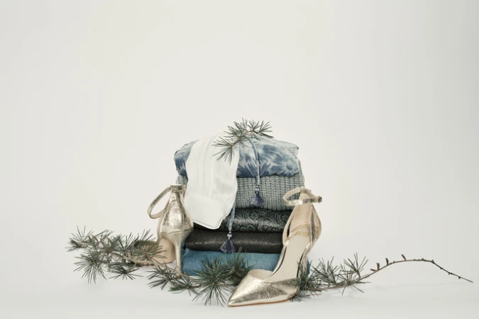 Stack of folded blue, white, and dark-colored fabrics, accessorized with gold high heels and evergreen branches on a white backdrop.