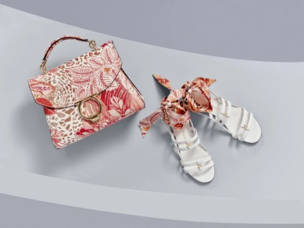 Fashion handbag with a floral print and white sandals with ribbon laces displayed on a light blue surface in a studio setting.