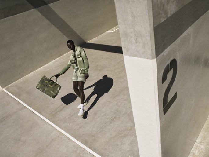 A dark-skinned adult model in a light-green matching jacket and shorts, holding a bag, stands in a concrete, architectural space.