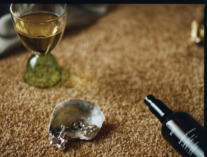 Still life featuring a glass of amber liquid, jewelry in a shell, and a cosmetic bottle on a textured brown surface, creating a muted, artistic composition.