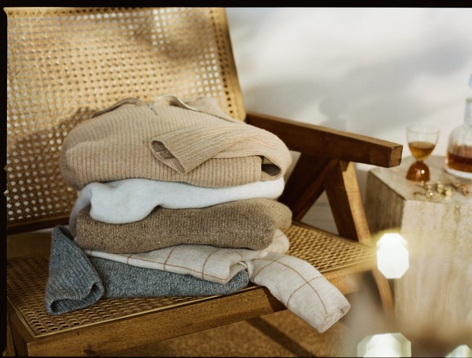 Stack of folded sweaters in neutral tones on a light brown wicker chair next to a stone pillar and glass liquor bottle indoors.