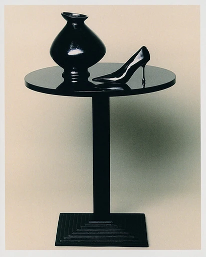 Black lacquered still life: a vase and stiletto heel pump are displayed on a round table with a square tiered base, against a neutral backdrop.