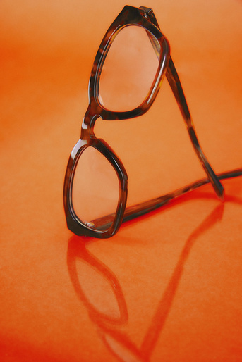 Eyeglasses with a tortoiseshell frame sit on a reflective bright orange surface, casting a soft shadow; studio lighting.