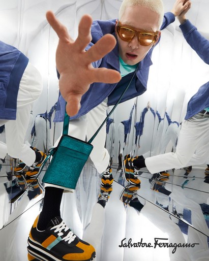 Fashion shot featuring an adult male model in a mirrored environment wearing sunglasses, a blue jacket, white pants, and black, orange, and white sneakers.
