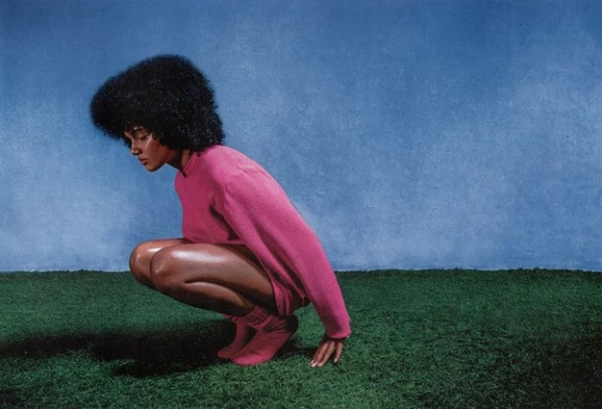 A woman with dark voluminous hair wearing a pink top and socks crouching on artificial green turf against a blue backdrop.