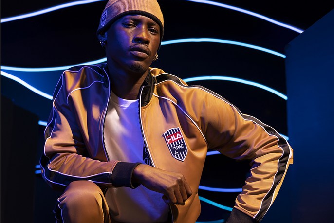 Portrait of a young Black man wearing a tan beanie and matching track suit, kneeling in a studio with blue neon lights in the background.