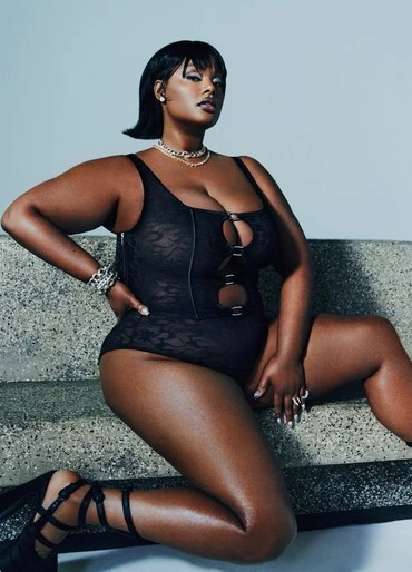 A dark-skinned adult female poses in a black lace bodysuit and high heels on a concrete surface, wearing jewelry and a blunt bob hairstyle.