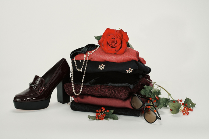 Stack of folded red and black sweaters and tops next to a red high-heeled shoe, with a red rose on top against white background.