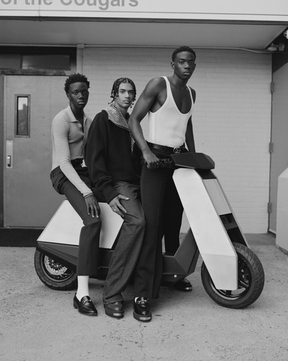 Three Black men in stylish casual wear pose with a futuristic white and black scooter against an indoor/outdoor urban backdrop.