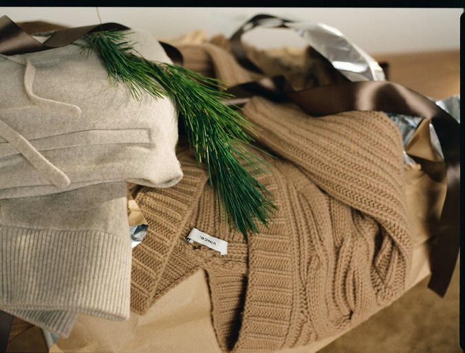 Beige and cream folded knitwear garments sit stacked and wrapped with ribbons and a small evergreen bough, suggesting a gift.