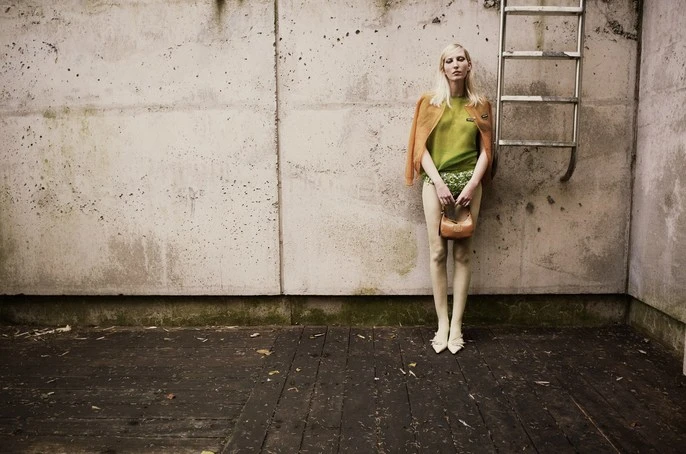 A blonde woman with an orange jacket poses in front of a textured concrete wall, holding a tan purse in an outdoor setting.