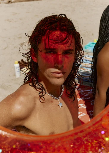 Young man with a silver chain and a star pendant is on the beach with a red-tinted face, joined by another person out of focus.