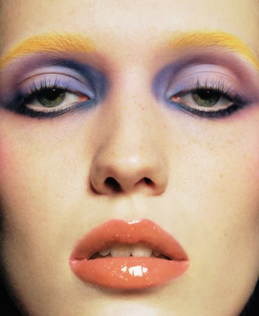 Close-up beauty portrait of a woman with vibrant yellow eyebrows, purple eyeshadow, and glossy coral lips by Olivia Giles