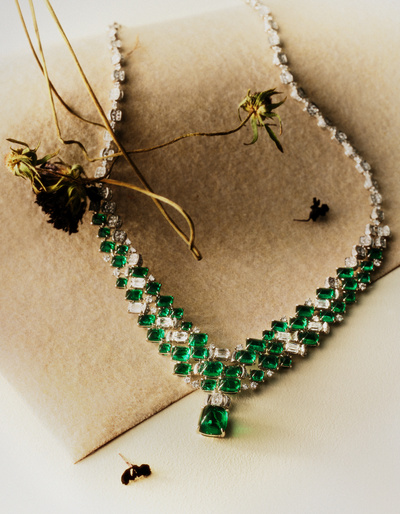 Elegant emerald and diamond necklace on beige fabric, complemented by dried floral accents in a softly lit, high-angle jewelry shot.