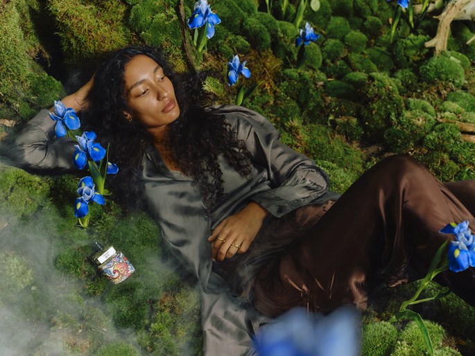 Woman in silky loungewear lying in a mossy, floral environment. An ornate cigarette case is beside her, adding a vintage touch.