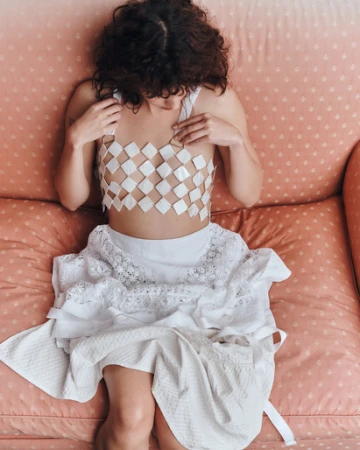 Model with dark curly hair wearing a white diamond-patterned top and layered white lace skirt seated on a pink patterned sofa