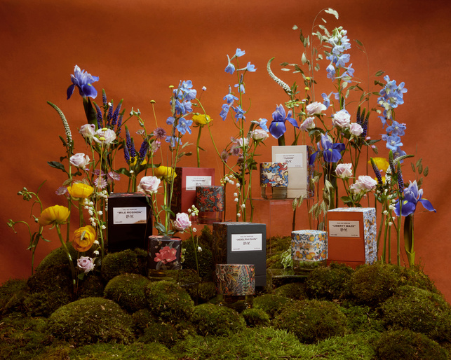 Still life of candle products on mossy platforms with blooming flowers against a solid orange backdrop, capturing beauty and fragrance.