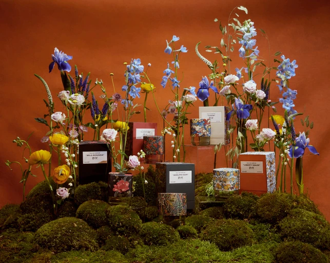 Still life of candle products on mossy platforms with blooming flowers against a solid orange backdrop, capturing beauty and fragrance.